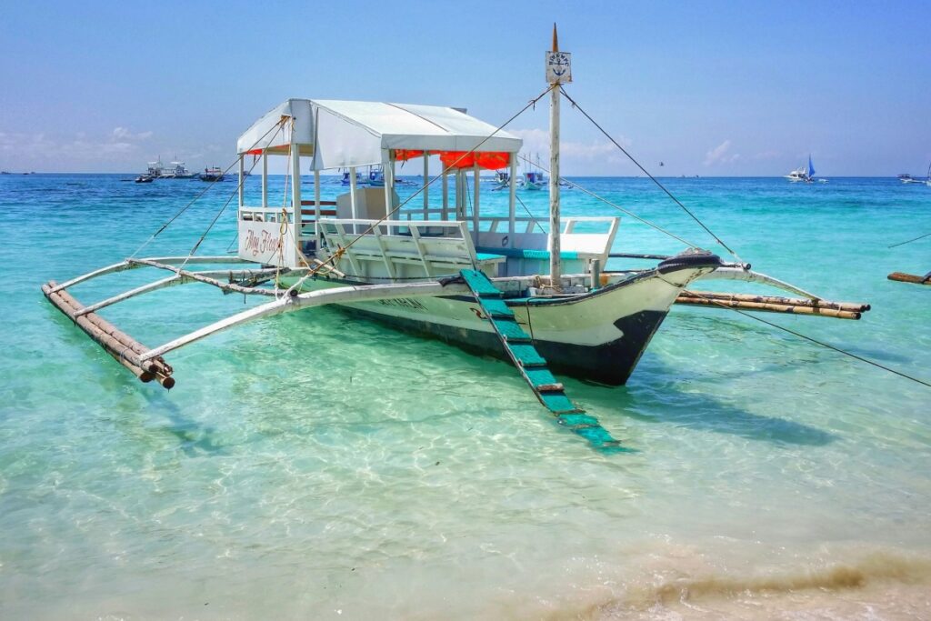 Isang bangka ng outrigger na nakapark sa malinaw at maputlaang tubig ng karagatan. Gabay sa Pagpili ng Tamang Influencer para sa Iyong Kampanya. Ang bangka ay may puting bubong at mga upuan, at may isang hagdanan na patungo sa loob nito. Sa likod, makikita ang iba pang mga bangka at ang karagatan na umaangat hanggang sa kalangitan.