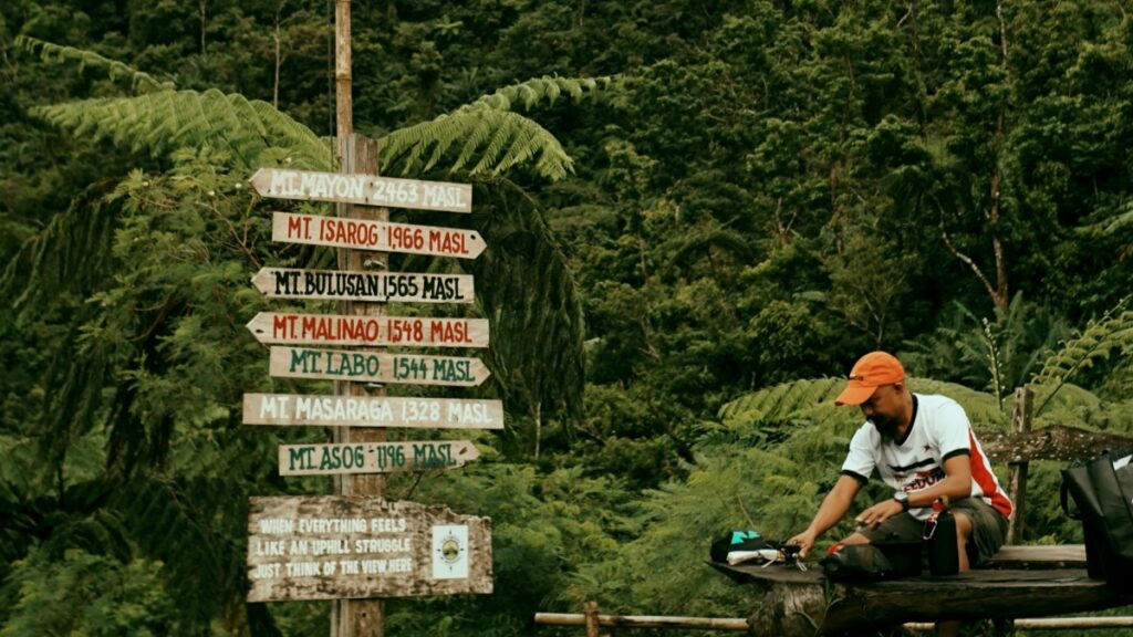 Ang larawan ay nagpapakita ng isang tao na nakaupo sa isang gubat na lugar kasama ang mga panandang patungo sa iba't ibang bundok sa Pilipinas. Kasama sa larawan ang mga pangalan ng mga bundok at taas nila. Samantala, sa blog post na "Paano Kumita ng Pera sa Bigo Live", tatalakayin kung paano gumana ang Bigo Live at mga hakbang sa pag-setup ng account.